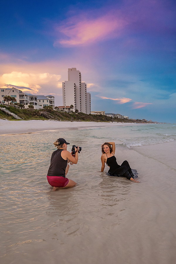 Seaside photographer behind the scenes capturing senior in black gown in surf at sunset