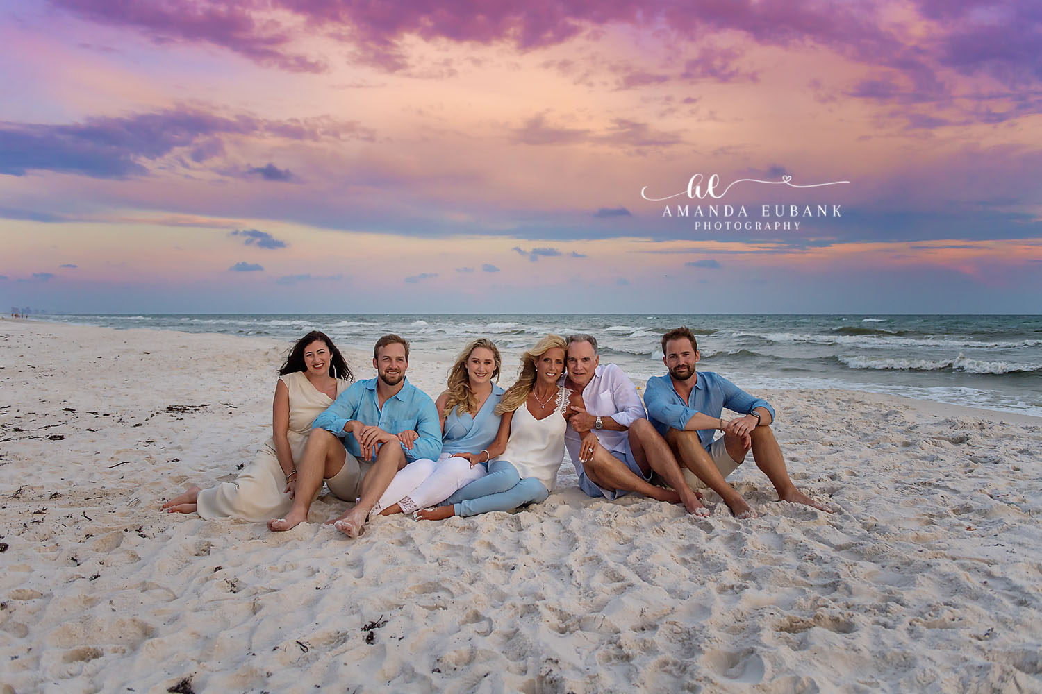 Seaside family photographer captures adult family of six sitting on white sand at pastel sunset