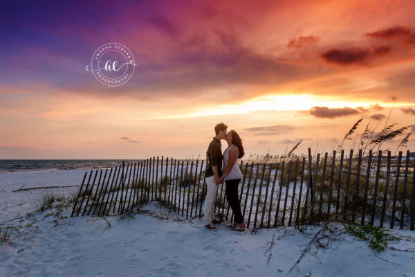 Seaside engagement photographer captures couple kissing by beach fence at dramatic sunset