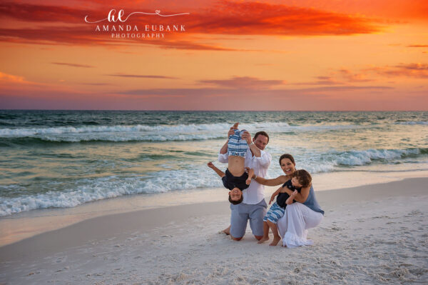 Seaside family photographer captures playful family with dad holding baby upside down at sunset