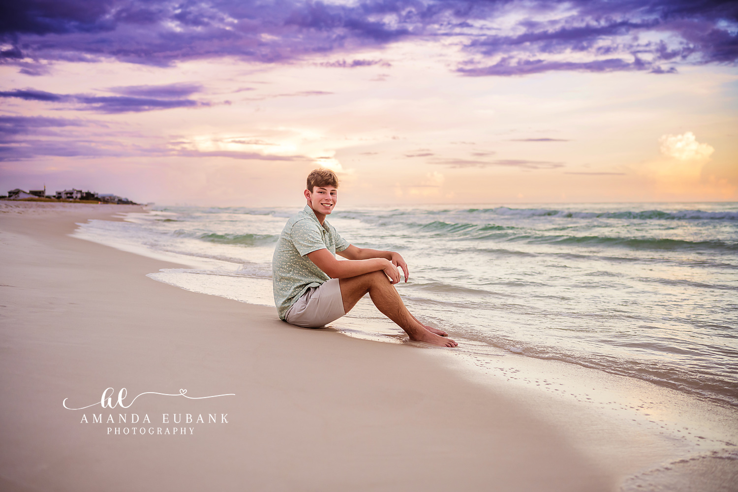 Seaside senior photographer captures male senior sitting at water edge in patterned polo at pastel sunset