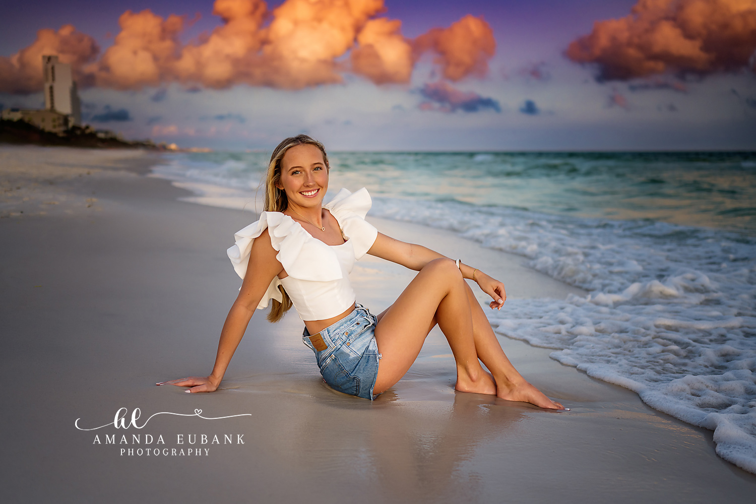 Seaside senior photographer captures blonde in white ruffle top and denim shorts sitting in surf at sunset