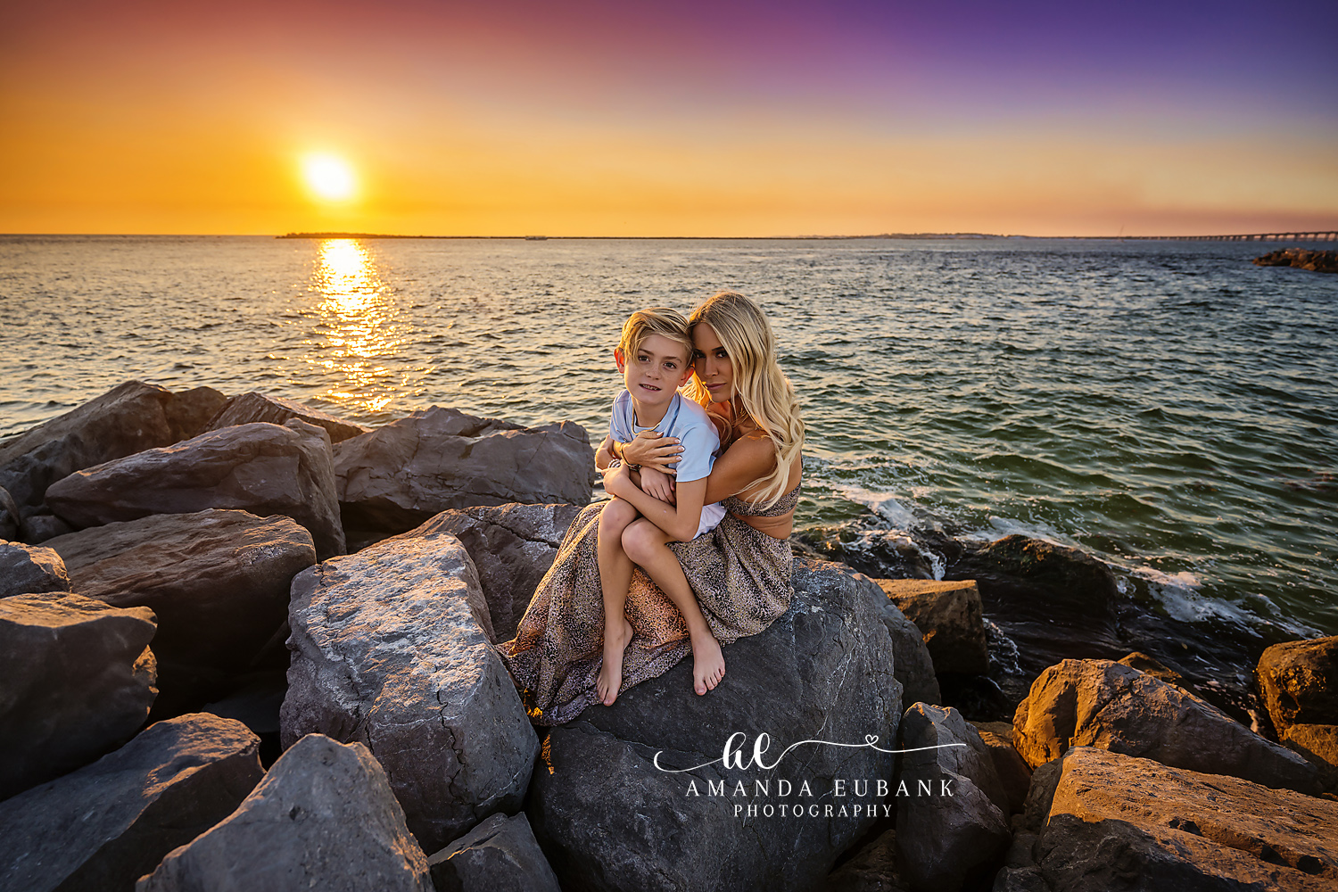 Seaside family photographer captures mom and son on rocky jetty at vibrant orange Bay sunset