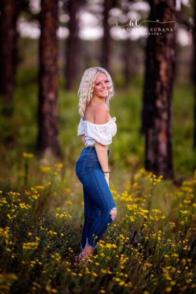 Seaside senior photographer captures blonde senior in white off-shoulder top in yellow wildflower field