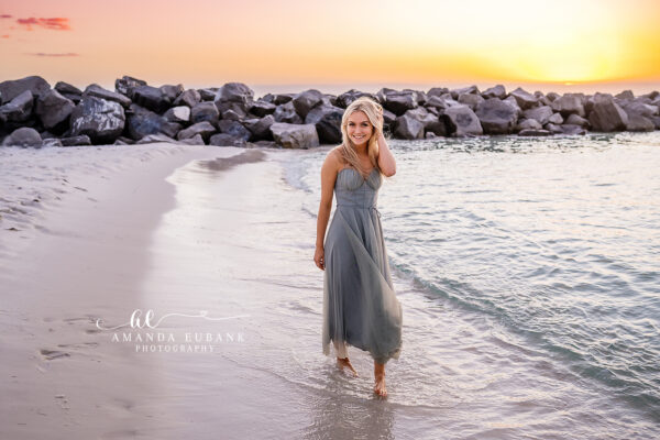 Seaside senior photographer captures blonde senior in gray gown wading along jetty rocks at sunset