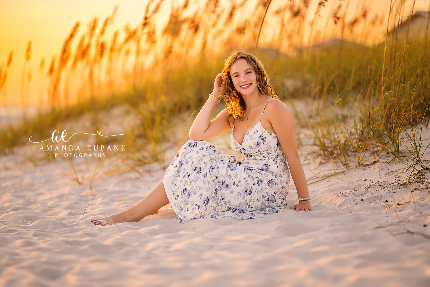 Seaside senior photographer captures curly-haired senior in floral dress sitting in sand among sea oats at sunset