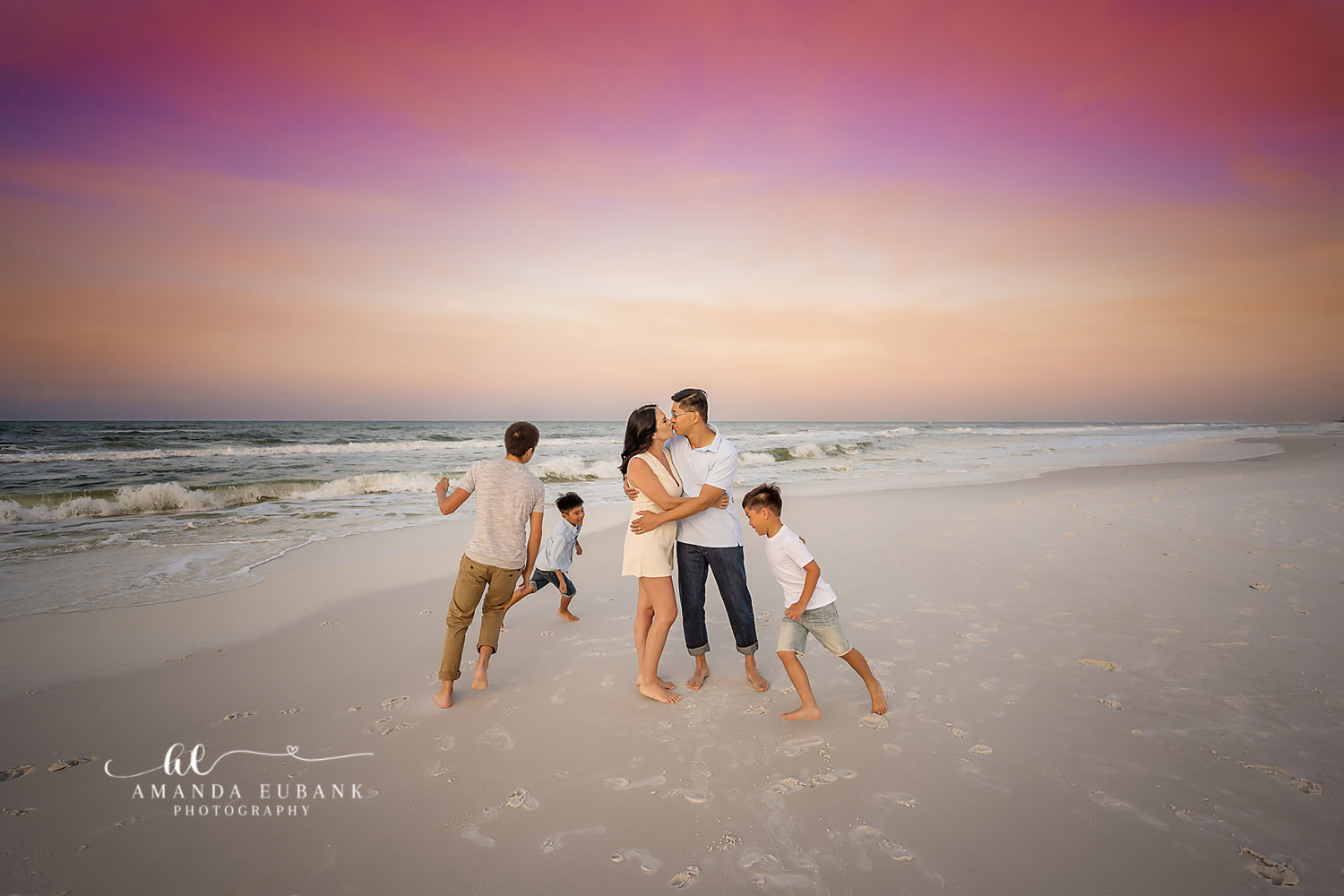 Seaside family photographer captures parents kissing while sons play around them at pink sunset